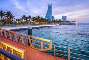 Beautiful daytime view of Jeddah, Saudi Arabia, showing modern waterfront buildings reflected on the calm Red Sea waters under clear skies, capturing the elegance of the city’s urban development.
