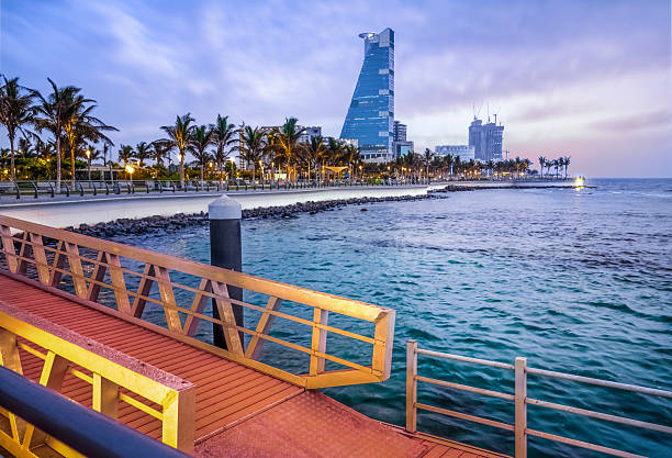 Beautiful daytime view of Jeddah, Saudi Arabia, showing modern waterfront buildings reflected on the calm Red Sea waters under clear skies, capturing the elegance of the city’s urban development.